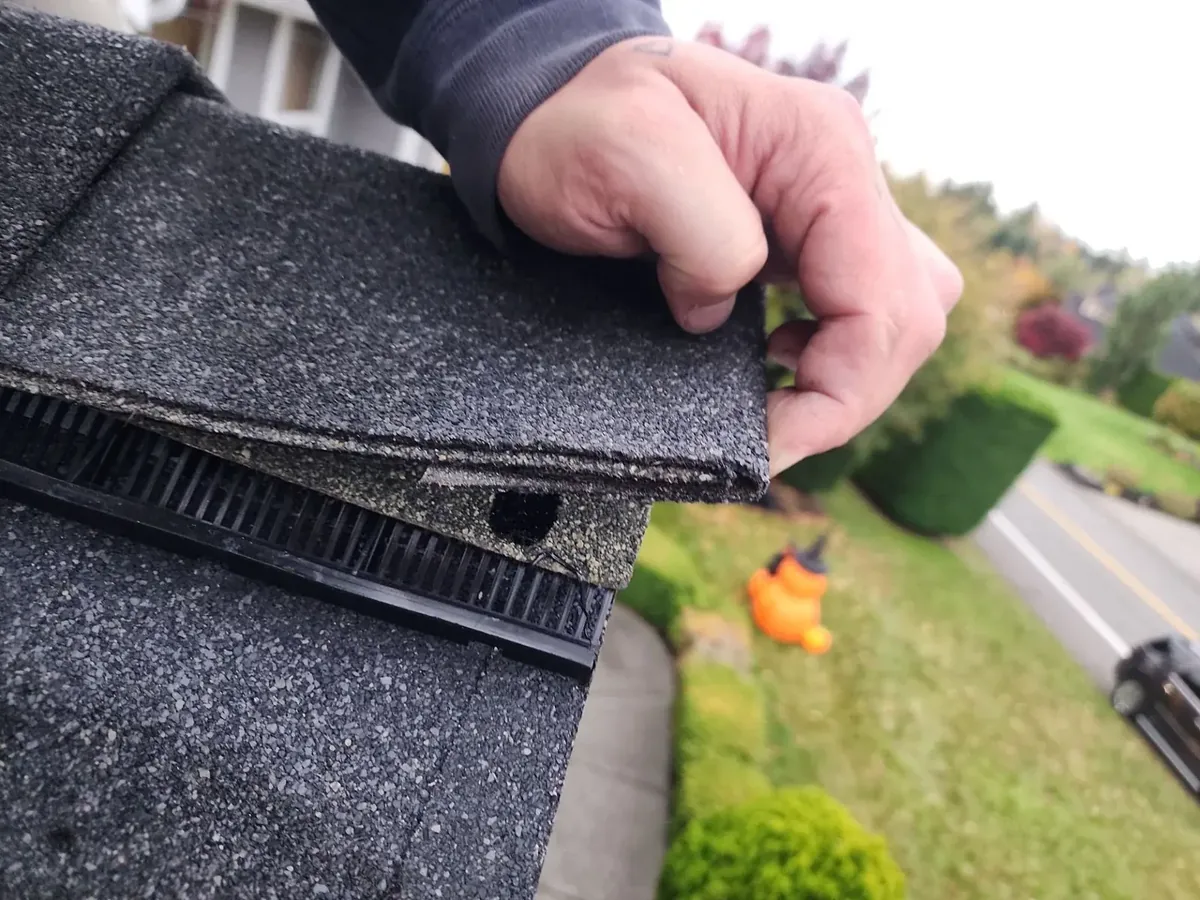 Hand lifting a shingle tab near the ridge to check its seal during roof inspections