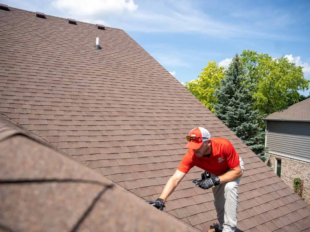 Roofer bending to check brown asphalt shingles on a steep roof during roof inspections