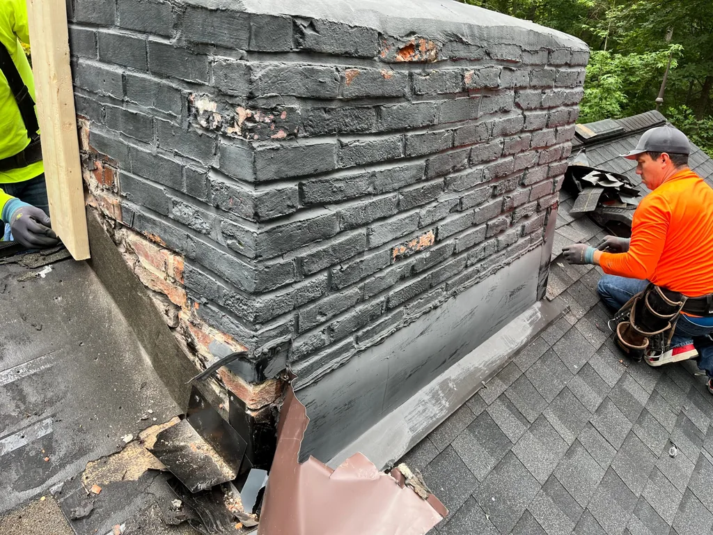 Two roofers tearing out old flashing at the base of a brick chimney for roof leak repair